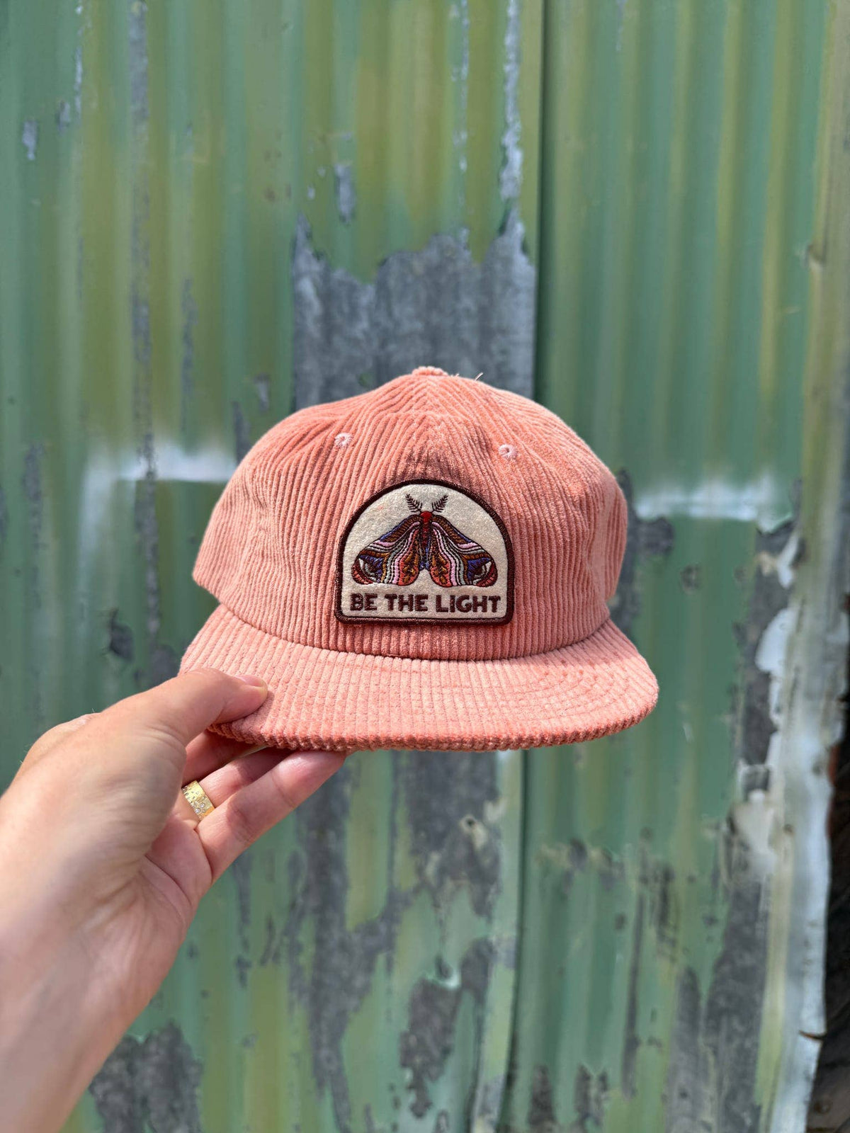 Hand holding the Be The Light corduroy cap in front of a green corrugated metal wall