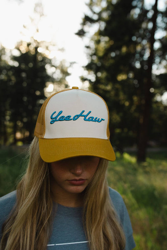 Yeehaw embroidered trucker hat in mustard gold and cream, worn by a woman with long blonde hair in a pine forest