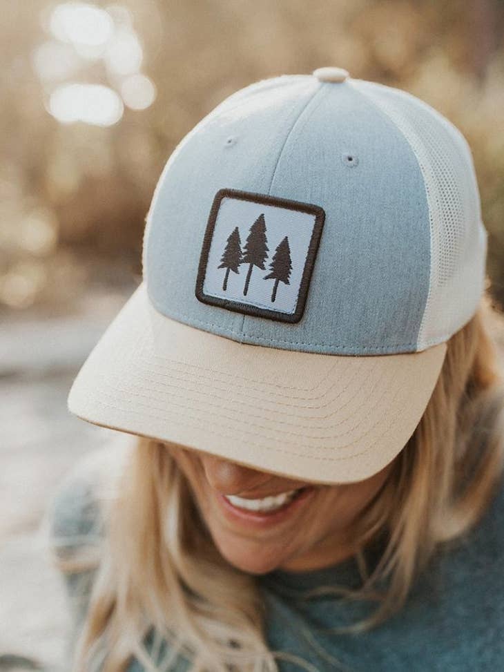 Woman in a flannel wearing the Three Tree tri-tone trucker hat, light gray-blue crown with a cream bill and three-pine patch
