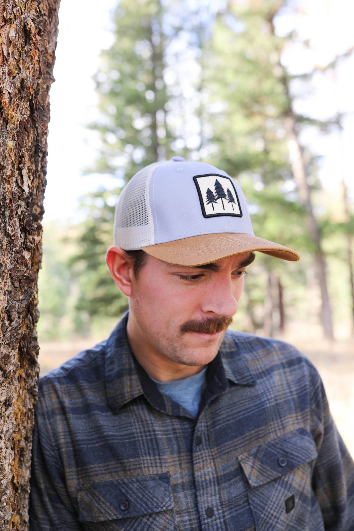 Man in a plaid flannel wearing the Three Tree trucker hat, leaning against a pine trunk