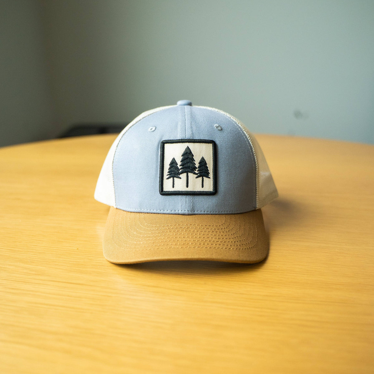 Three Tree tri-tone trucker hat on a yellow table, gray crown with a mustard bill and three-pine embroidered patch