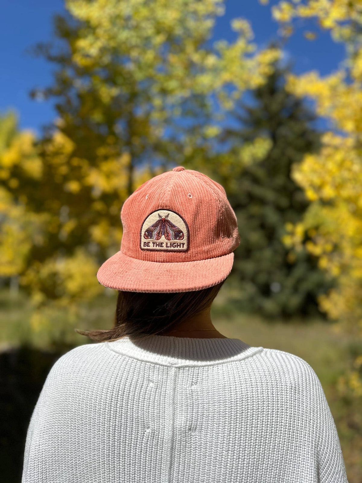 Person in a cream waffle-knit top seen from behind wearing the Be The Light corduroy cap among autumn trees