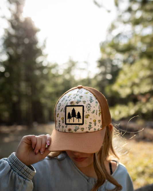 Three Tree floral trucker hat with a three-pine patch on a wildflower print panel, worn by a woman tipping the brim outdoors