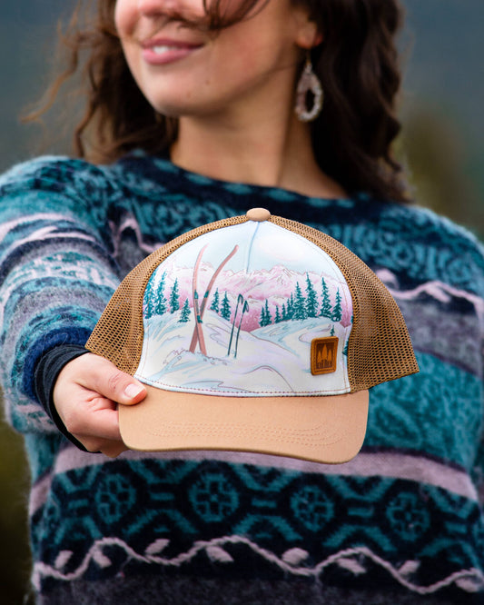 Moonlight Ski trucker hat with a pastel snowy ski-slope front panel and light brown mesh back, held out by a woman in a patterned sweater