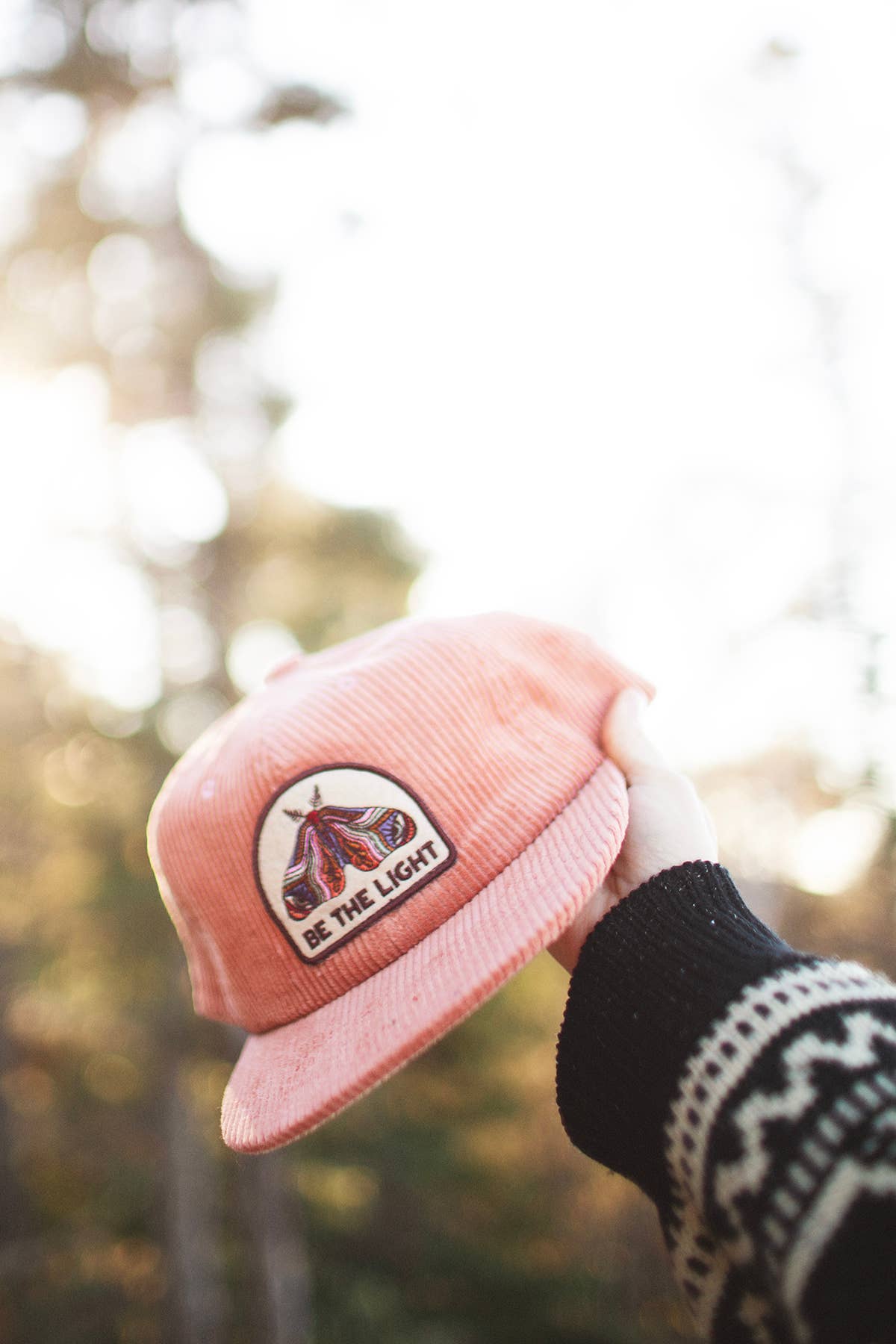 Hand in a patterned sweater holding up the Be The Light corduroy cap in front of pine trees