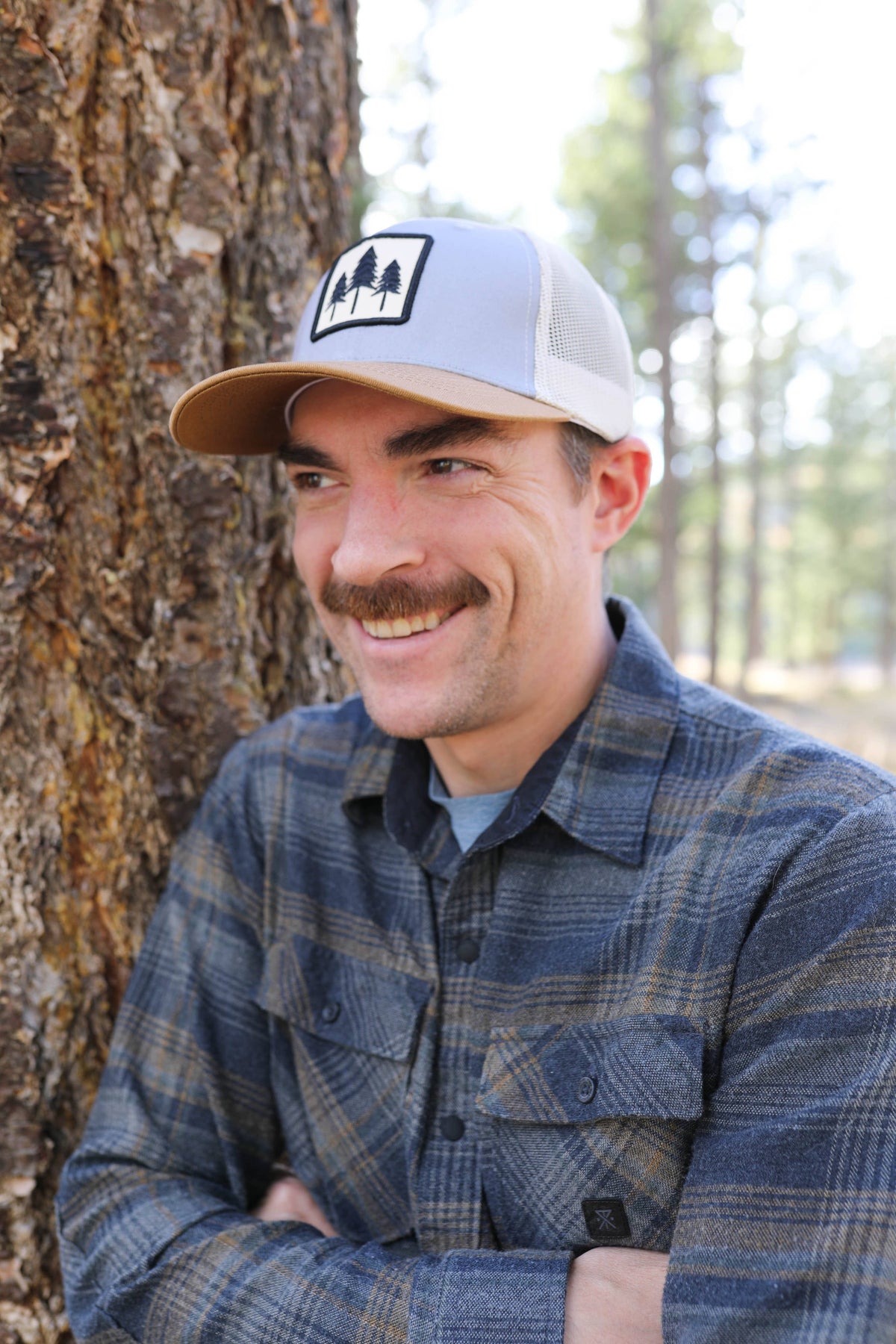 Man in a plaid flannel wearing the Three Tree tri-tone trucker hat in a pine forest